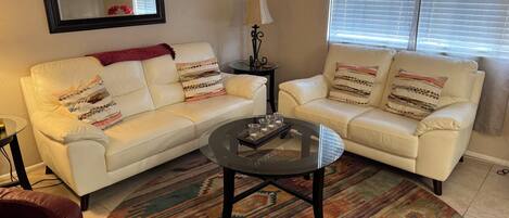 A living room with two white leather sofas, a glass coffee table, a patterned rug, a lamp, and a television on the wall.