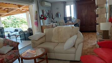 Un salon avec un canapé beige, une petite table ronde et un fauteuil rouge. On y trouve également un coin cuisine avec une table à manger et des chaises, ainsi qu’une grande fenêtre avec des rideaux.