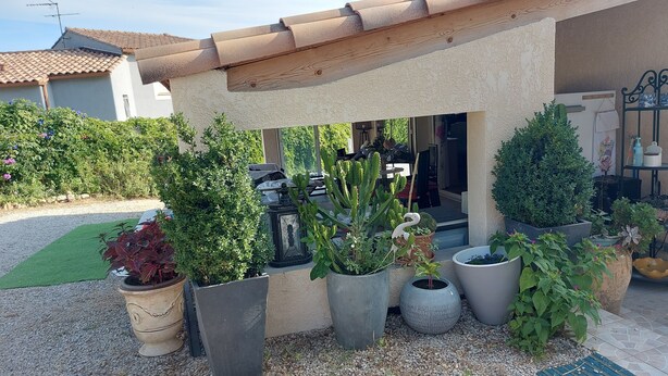 Un espace extérieur abrité, agrémenté de diverses plantes en pot, d’un chemin de gravier et d’où l’on aperçoit une maison au loin.