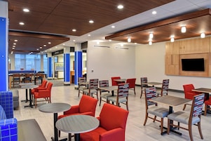 A modern dining area with multiple tables and chairs, a flat-screen TV mounted on the wall, and a view of the outdoors through large windows.