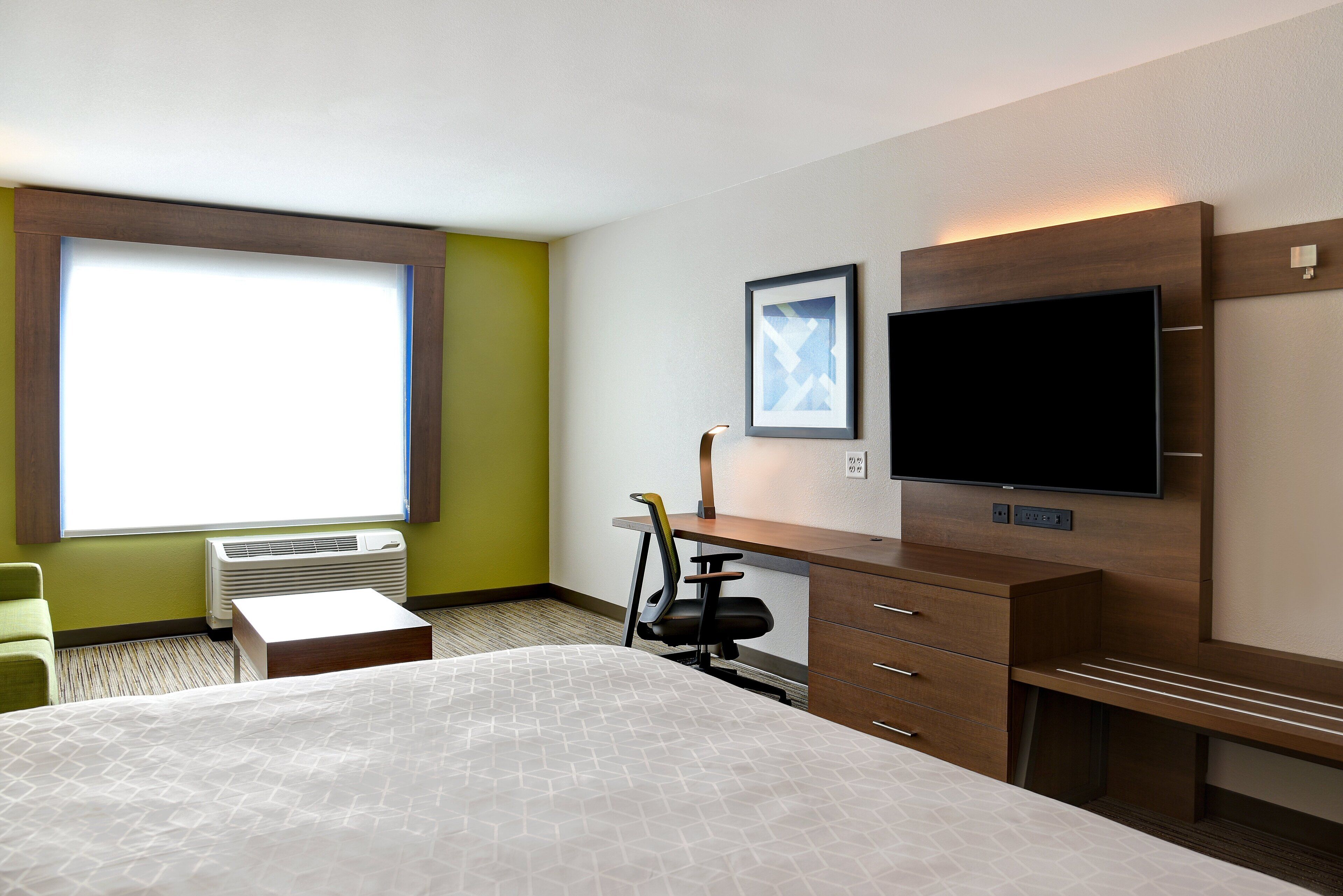 A hotel room with a bed, desk, chair, TV, and air conditioning unit.