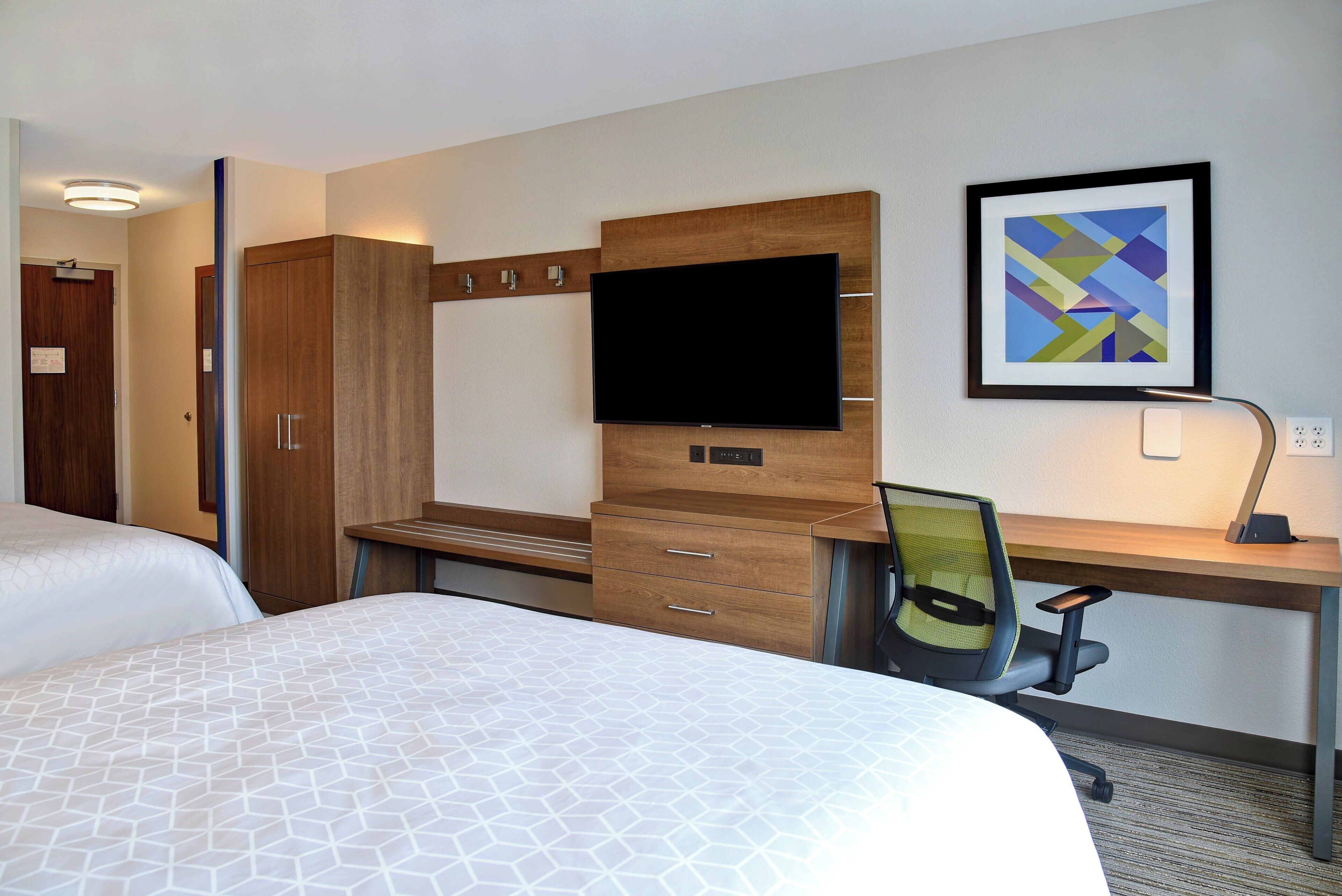 A hotel room with a bed, a desk with a chair, a TV, and a framed picture on the wall.