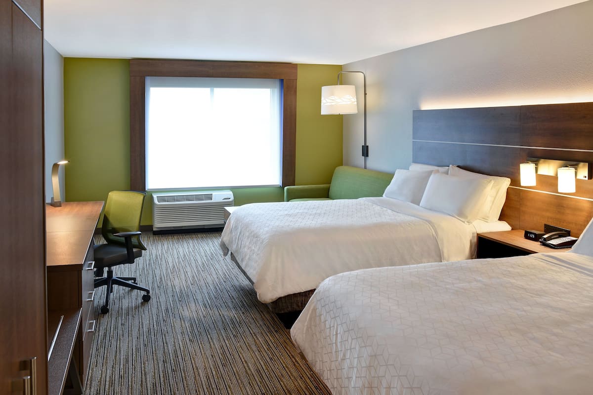 A hotel room with two beds, a desk, a chair, a lamp, and a window.