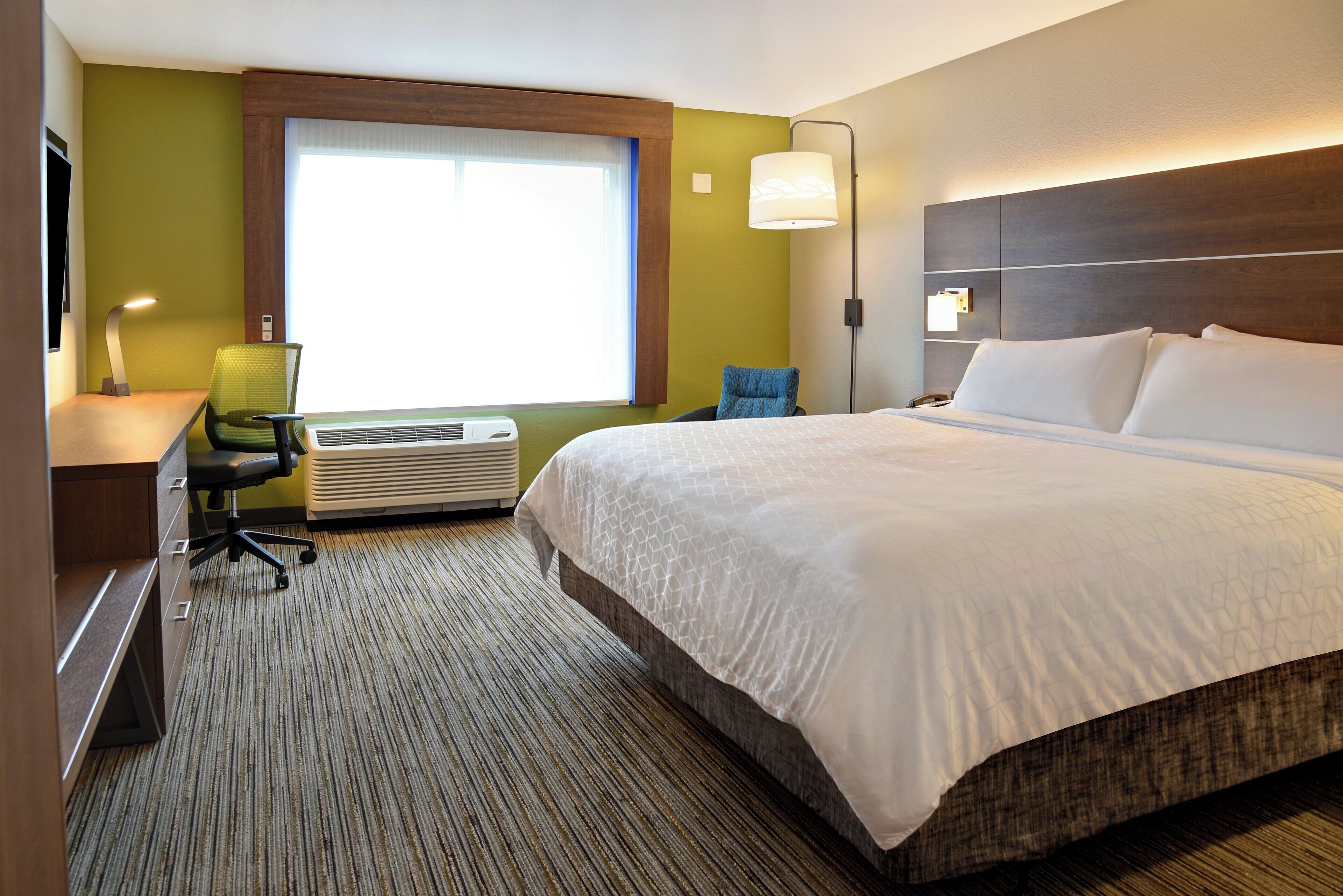 Quarto de hotel com uma cama grande, uma escrivaninha com cadeira, uma janela com persianas e uma luminária na parede.