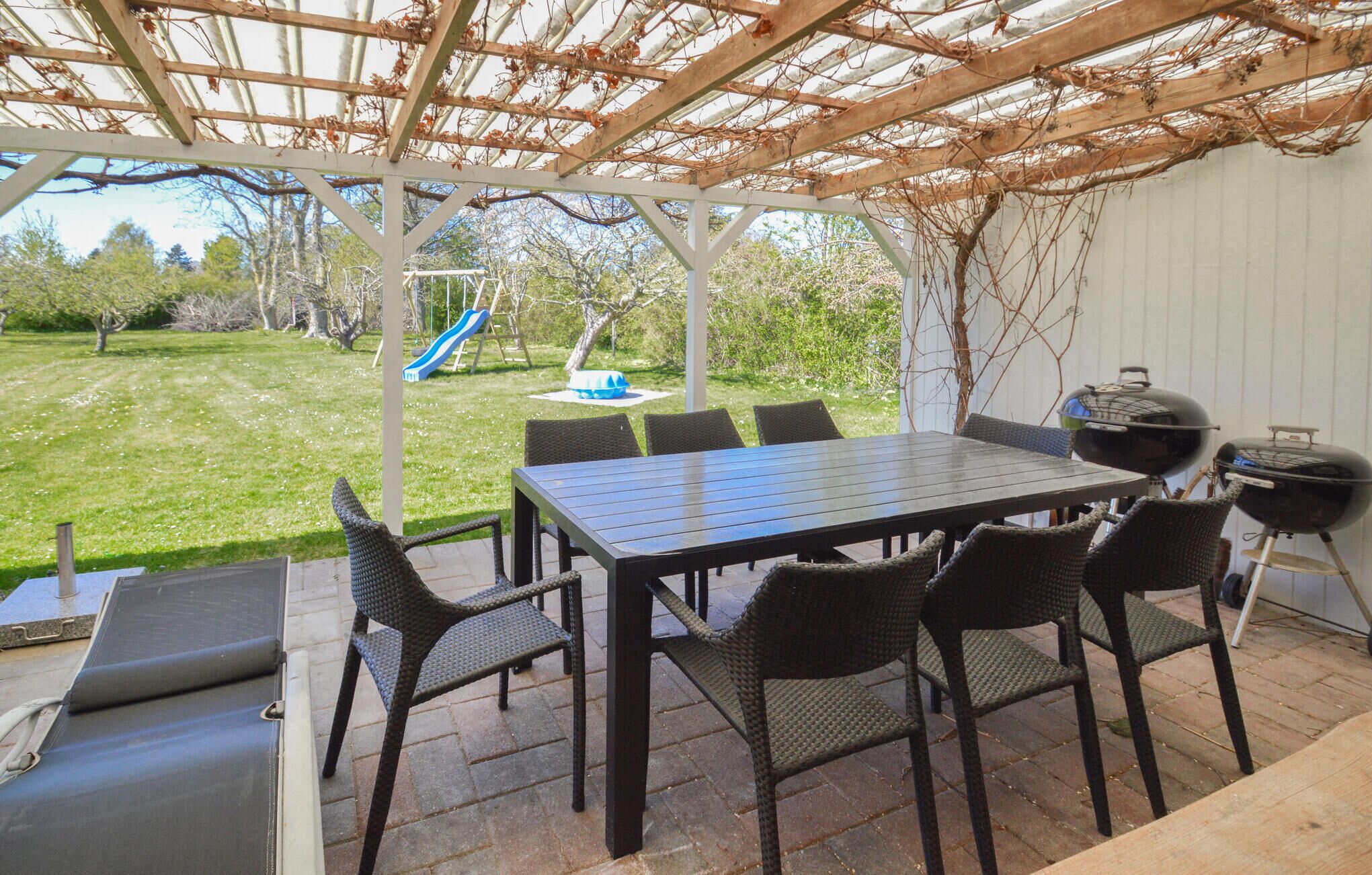 En overdækket terrasse med et bord og stole, et gyngestativ og en grill.