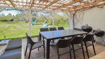 En overdækket terrasse med et bord og stole, et gyngestativ og en grill.