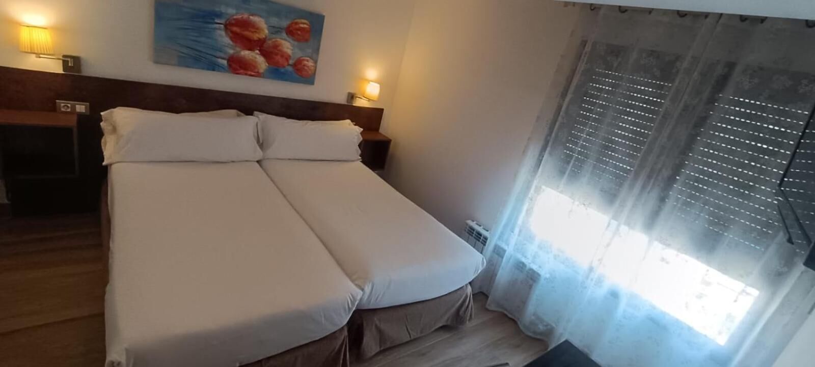 Una cama con sábanas blancas, cabecera, un cuadro en la pared y una ventana con persianas.