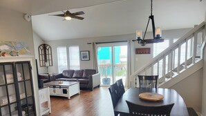 A spacious living room with a dark brown leather sofa, a white coffee table, and a dining table with chairs. There is a staircase with white railings leading to a balcony with a view of the outside. - Jamaica Beach! Fishing light, kayaks, pool access! (Jamaica Beach)