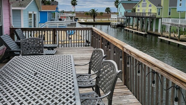 A wooden deck with a table and chairs, overlooking a water body with colorful buildings in the background. - Jamaica Beach! Fishing light, kayaks, pool access! (Jamaica Beach)
