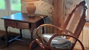 A rustic room with a wooden rocking chair, a small wooden table with a lamp, and a stone wall.
