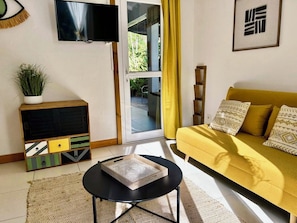 A living room with a sofa, a coffee table, a TV, a plant, and a sliding glass door. - Atemoya lodges. Swimming pool, beach 400 m., Cinnamon (4p), A/C, pool, beautiful garden, beach 400 m (Deshaies)