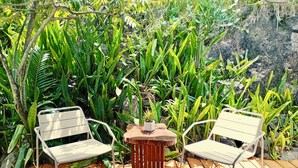 Deux chaises en métal blanc, séparées par une petite table ronde, sont placées sur une terrasse en bois, elle-même entourée d’un luxuriant feuillage verdoyant.
