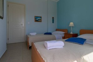 Quarto de hotel com duas camas, uma porta, uma mesinha de cabeceira, um abajur e duas pinturas penduradas na parede.