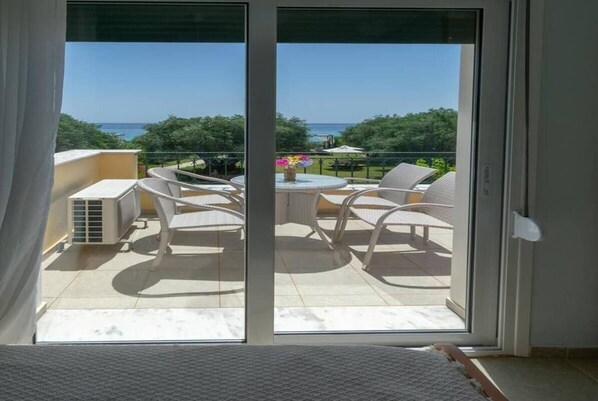 Uma varanda com mesa e cadeiras, um pequeno aquecedor e vista para o mar e vegetação.