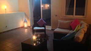 Een gezellige zithoek met een bank, een fauteuil en een salontafel. Er is een raam met gordijnen en een deur naar een balkon.