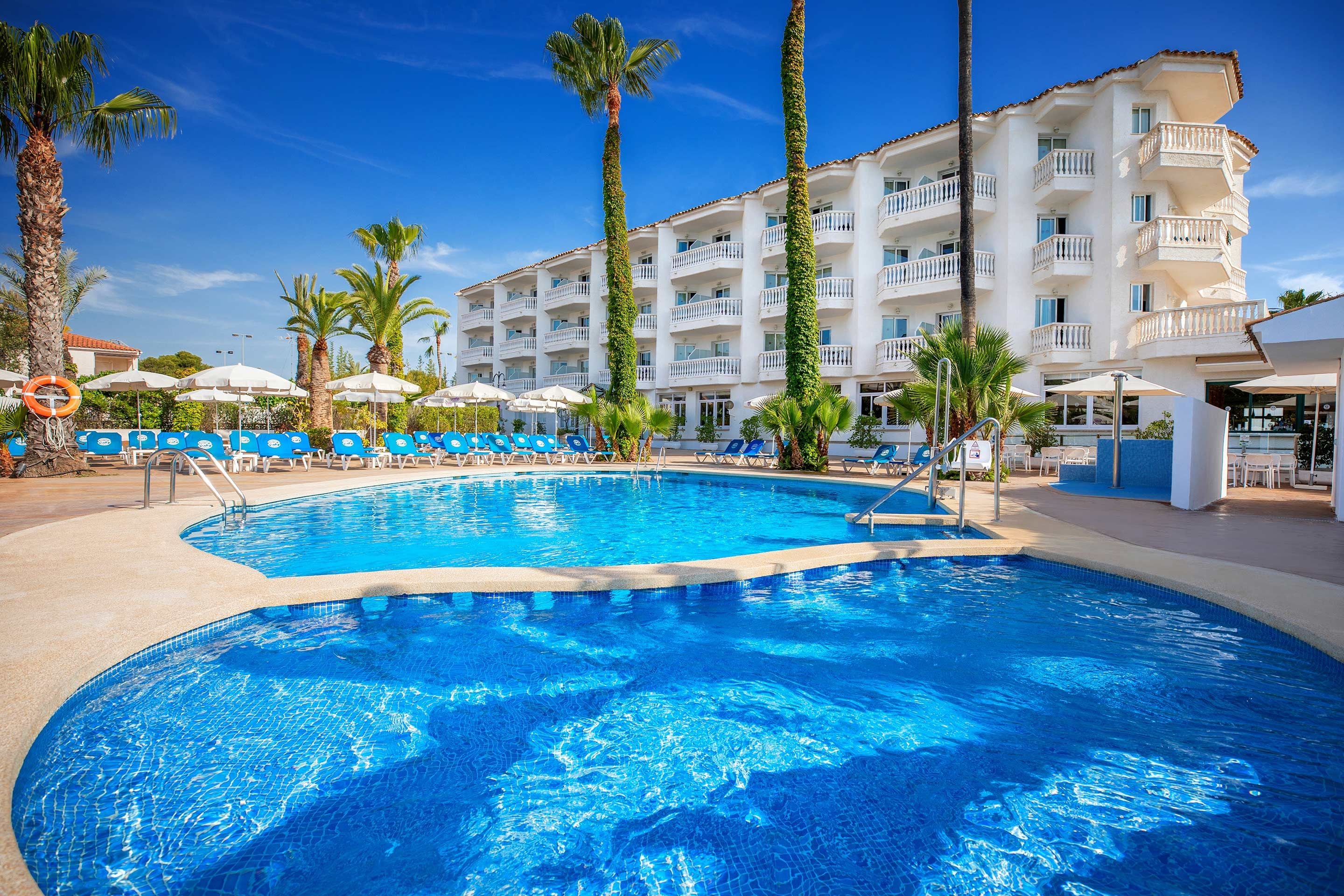 Una piscina con sillones de descanso, palmeras y un edificio de hotel de varios pisos.