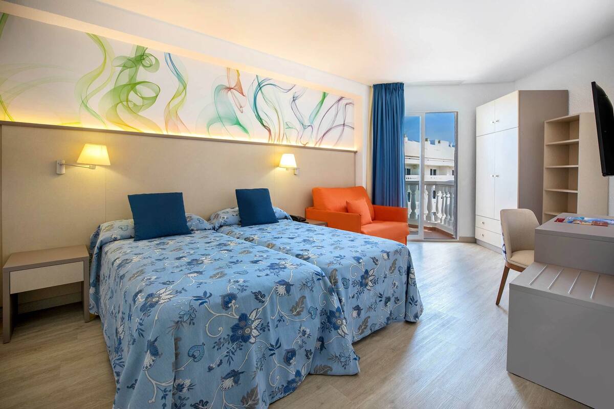 Une chambre d’hôtel avec deux lits, un bureau, une chaise, une armoire et une grande fenêtre donnant sur un beau paysage.