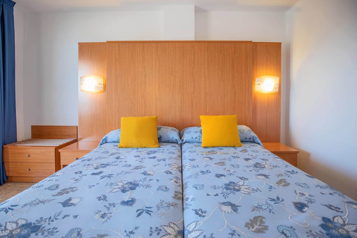Un lit double avec une literie à motifs floraux, deux tables de chevet avec des lampes, une tête de lit en bois et une petite commode en bois.