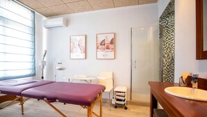 Une pièce comprenant une table de massage, une petite chaise blanche, une coiffeuse en bois avec un lavabo et une cabine de douche en verre.