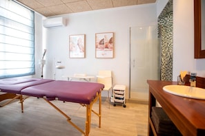 Ein Zimmer mit Massagetisch, einem kleinen weißen Stuhl, einem Holzanfangstisch mit Waschbecken und einer Glasdusche.