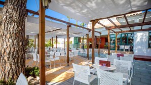 Un espace extérieur aménagé en terrasse, avec des chaises et des tables en métal blanc, un arbre, et une partie abritée d’où l’on peut admirer la rue.