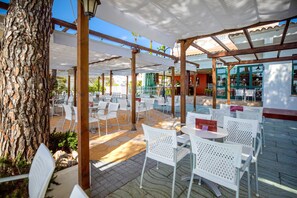 Ein Außenbereich mit einer Terrasse, weiße Metallstühle und -tische, ein Baum und ein überdachter Bereich mit Blick auf die Straße.