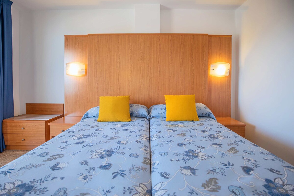 Ein Doppelbett mit floralem Bettbezug, zwei Nachttische mit Lampen, ein hölzernes Kopfteil und eine kleine Holzkommode.