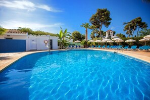 A swimming pool with lounge chairs and umbrellas, surrounded by trees and a building with a tiled roof. - Hotel Servigroup Romana (Alcala de Xivert)