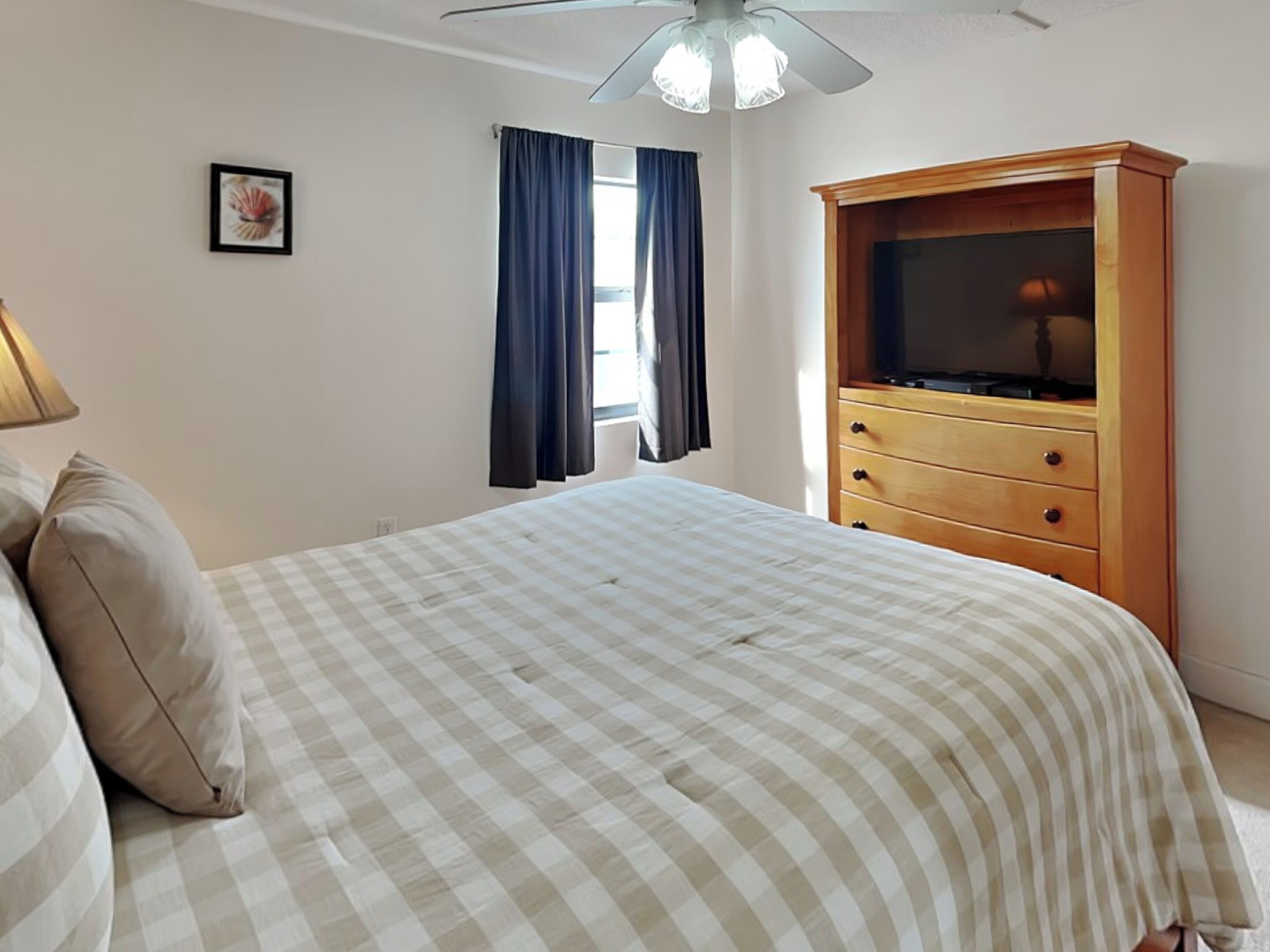 Een slaapkamer met een bed, een houten dressoir, een plafondventilator en een raam met gordijnen.
