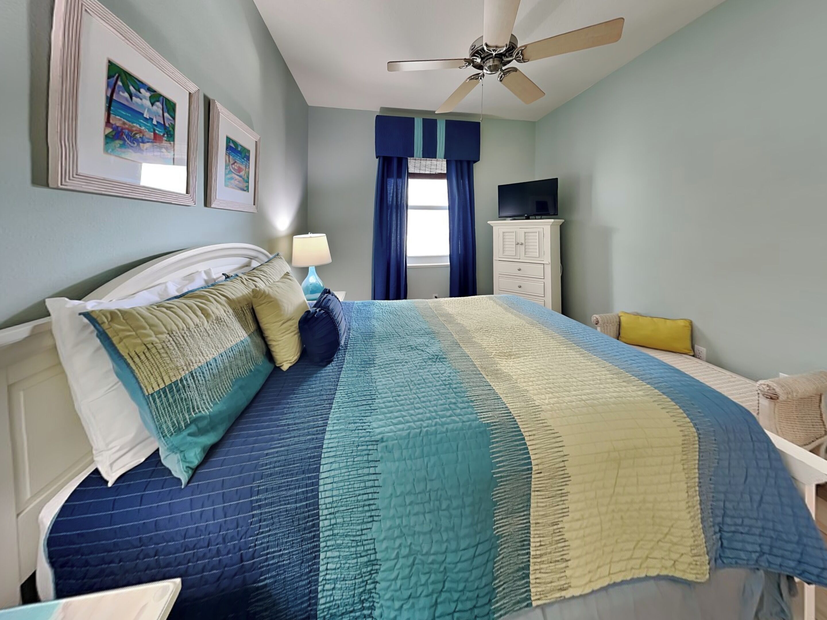 A bedroom with a bed, a ceiling fan, a TV, and artwork on the wall.