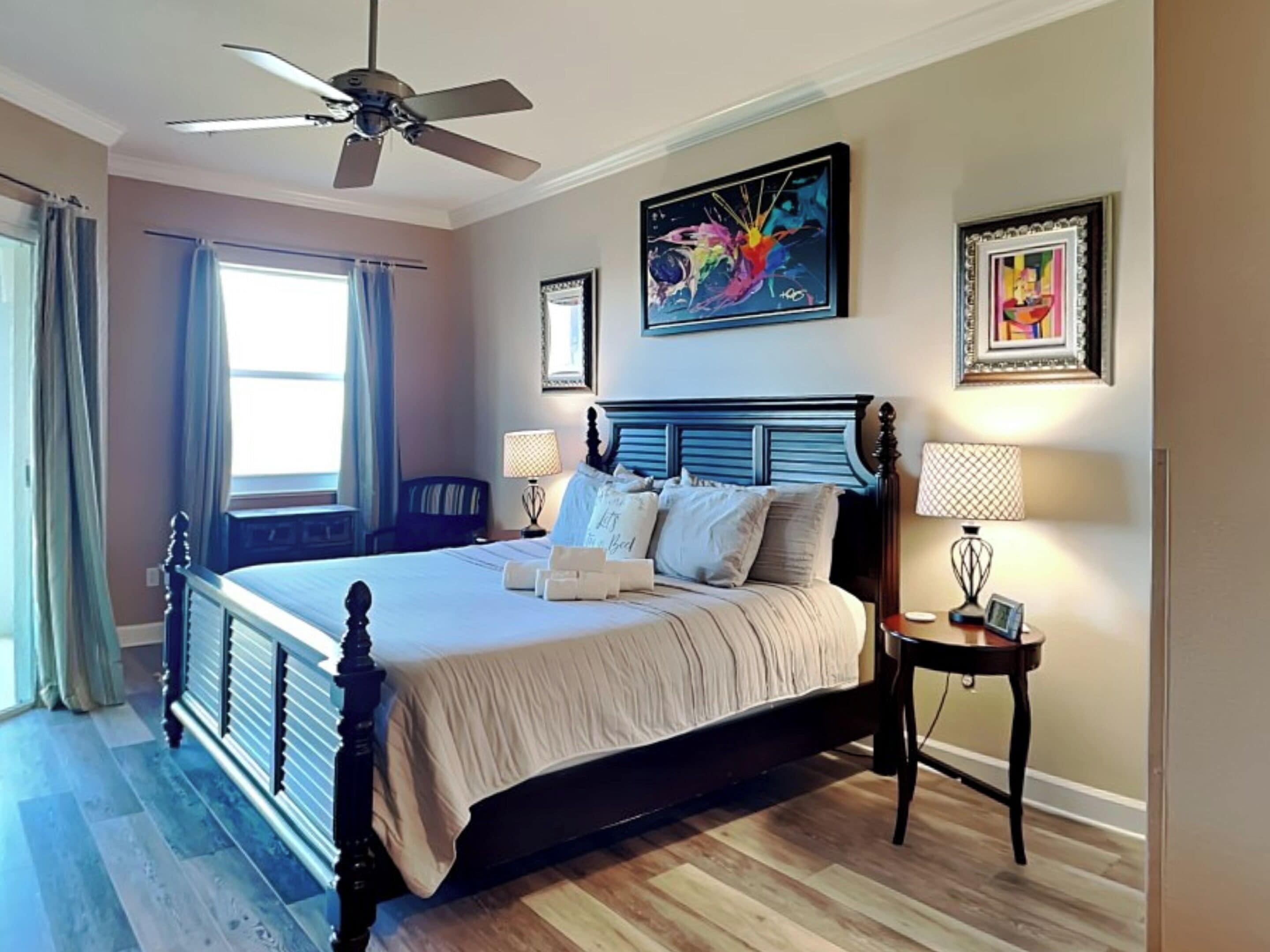 Une chambre avec un grand lit, deux tables de chevet, un ventilateur de plafond et des œuvres d’art accrochées aux murs.