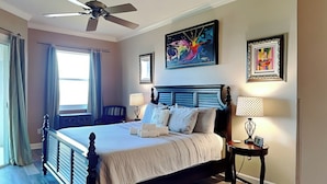 A bedroom with a large bed, two bedside tables, a ceiling fan, and artwork on the walls. - Cinnamon Beach 4 by Distinctive Beach Rentals (Palm Coast)
