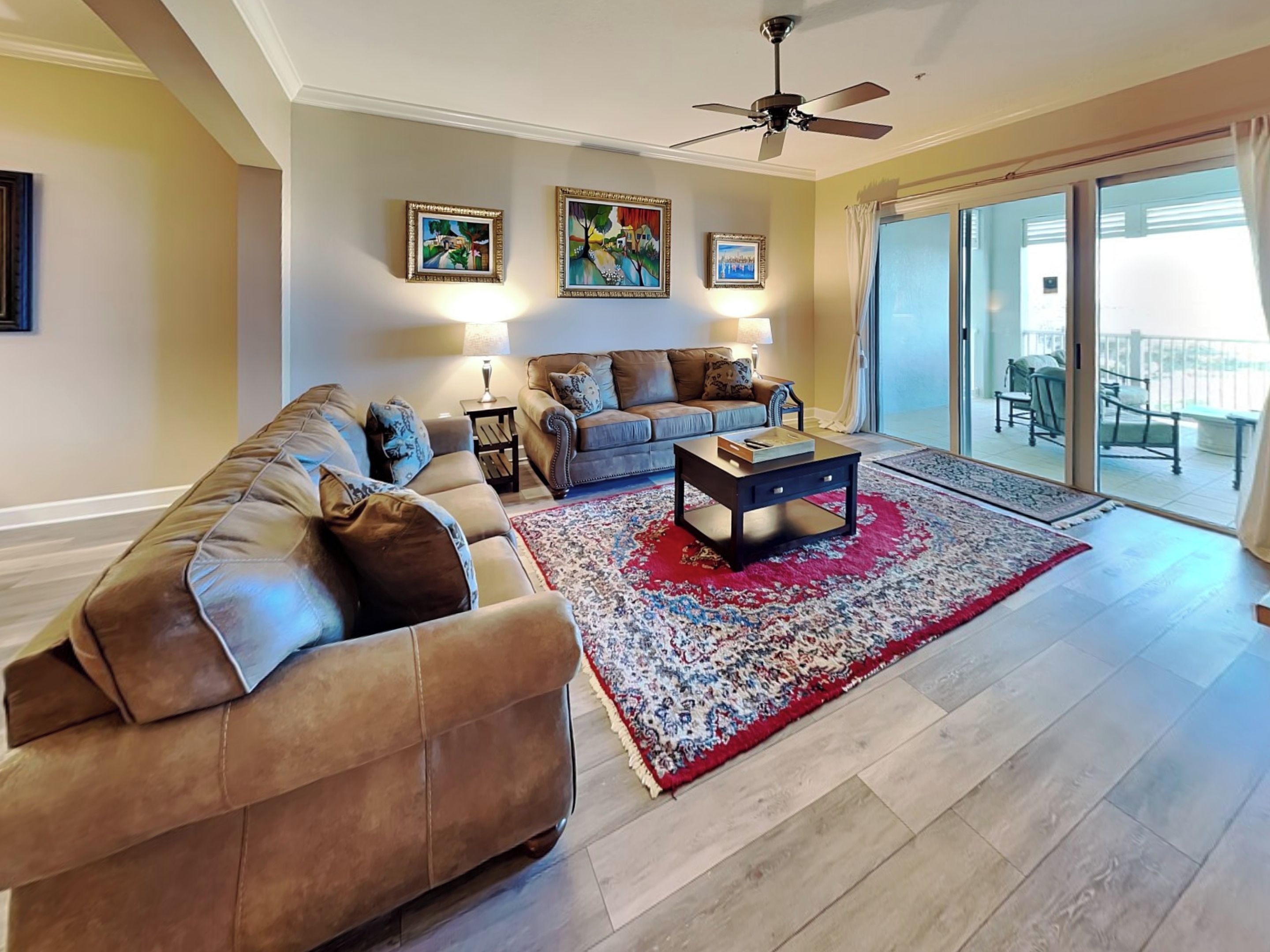 A spacious living room with a brown leather sofa, a blue sofa, and a coffee table. There are three framed artworks on the wall, and a ceiling fan is visible.