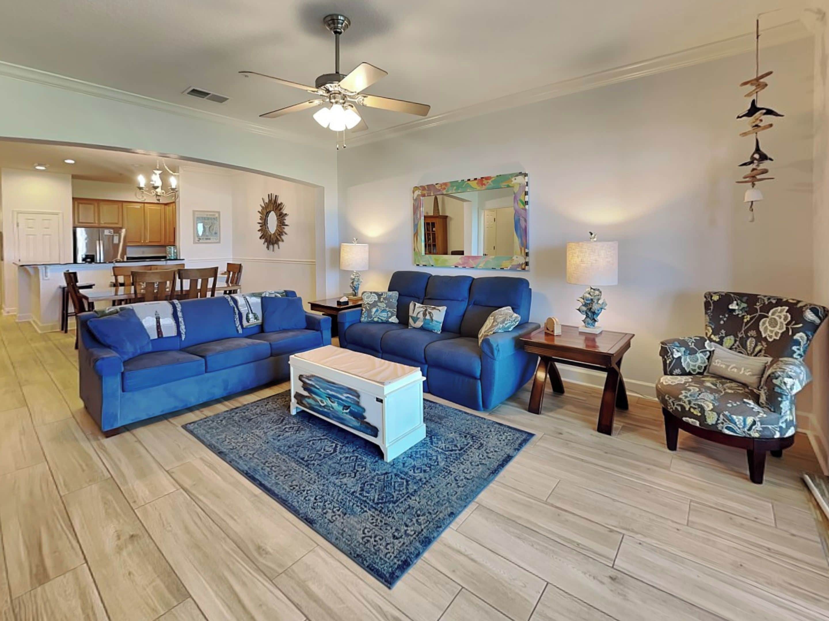 A living room with a blue sofa set, a coffee table, a patterned rug, a wooden floor, a ceiling fan, and a kitchen in the background.