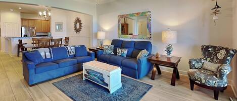 A living room with a blue sofa set, a coffee table, a patterned rug, a wooden floor, a ceiling fan, and a kitchen in the background.