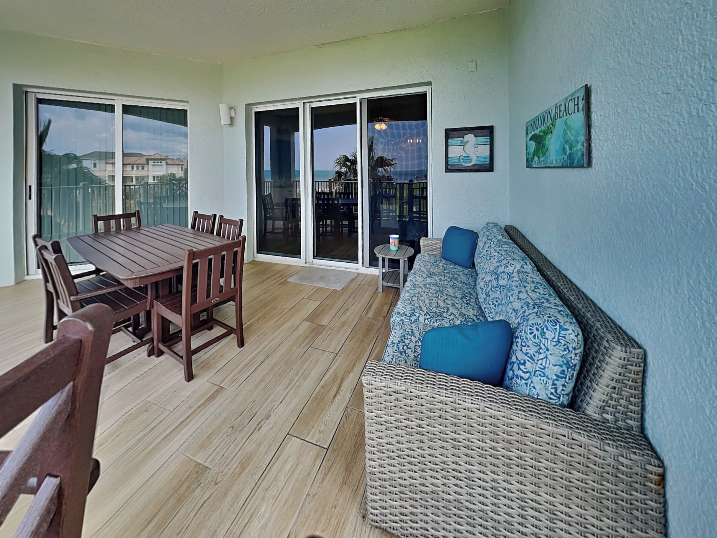 A spacious patio with a wooden table and chairs, a wicker sofa with blue cushions, and a sliding glass door leading to a balcony.