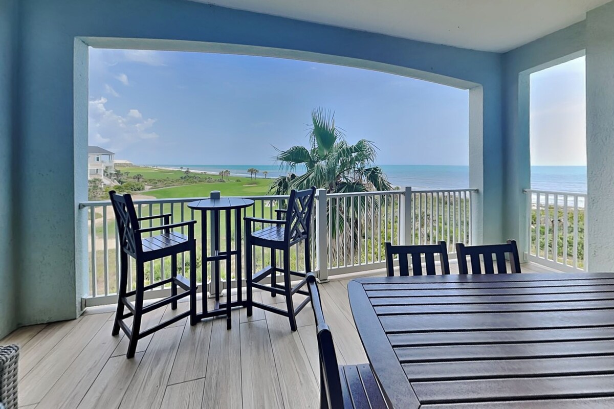 Een balkon met houten vloer, een tafel en vier stoelen. Het uitzicht is op een golfbaan en de oceaan.