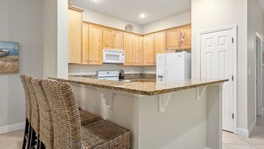 A kitchen with wooden cabinets, a granite countertop, a microwave, a refrigerator, and a wicker chair. - Cinnamon Beach 4 by Distinctive Beach Rentals (Palm Coast)