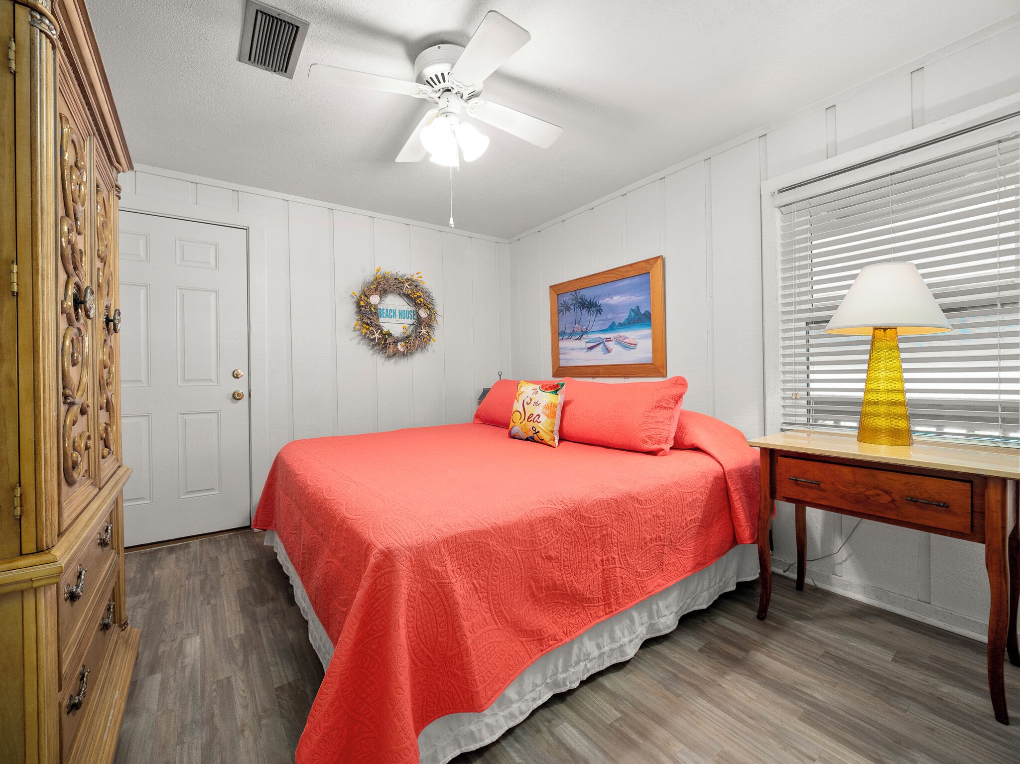 A bedroom with a bed, a desk with a lamp, a dresser, a ceiling fan, a door, a wall with a wreath, and a painting.