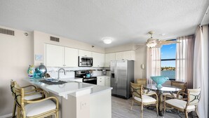 A modern kitchen with white cabinets, stainless steel appliances, a central island, and wicker chairs. - Casa Del Mar G-608 by Distinctive Beach Rentals (St. Petersburg)