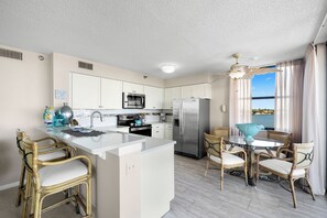 A modern kitchen with white cabinets, stainless steel appliances, a central island, and wicker chairs. - Casa Del Mar G-608 by Distinctive Beach Rentals (St. Petersburg)
