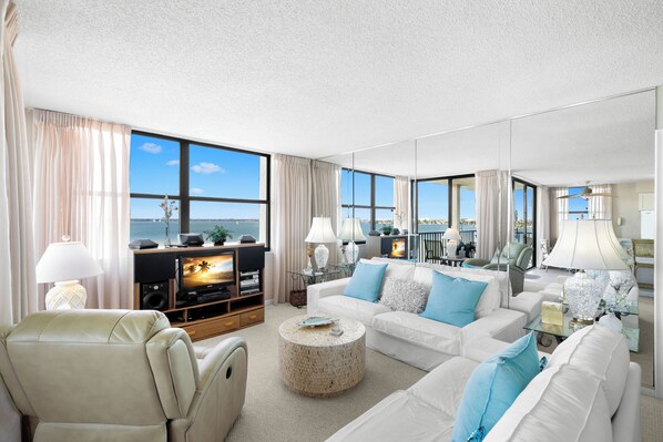 A spacious living room with a large window, a flat-screen TV, and a comfortable seating area. - Casa Del Mar G-608 by Distinctive Beach Rentals (St. Petersburg)