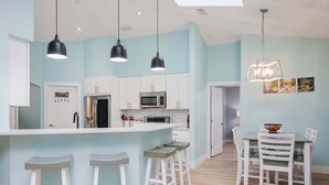 A modern kitchen with a central island, bar stools, and a dining area with white chairs. - Beach Daze by Distinctive Beach Rentals (Palm Coast)