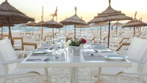Mesa de comedor frente a la playa con sillas blancas, un jarrón de flores y vista al océano.
