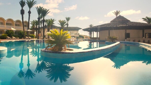Una piscina con palmeras, una estructura de techo de paja y un edificio con ventanas arqueadas.
