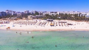 Una playa con filas de tumbonas y sombrillas, un cielo azul despejado y edificios al fondo.