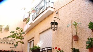 Un patio al aire libre con mesas y sillas de metal, plantas en macetas y un pequeño balcón con barandilla.