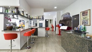Un moderno hall de hotel con un mostrador de recepción, taburetes rojos de bar, una zona de bar con varias botellas y un mostrador de recepción con un cartel.