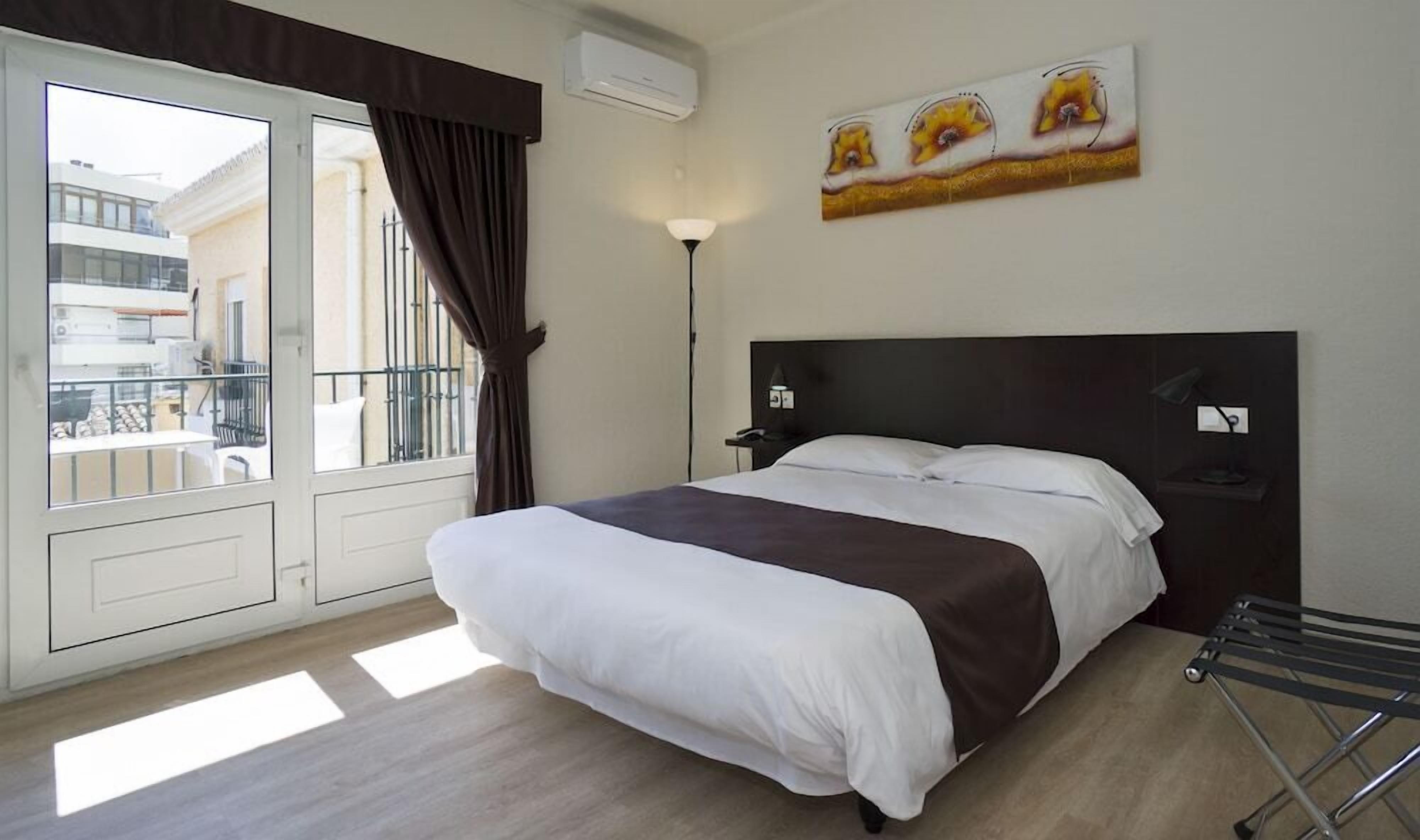 Ein Hotelzimmer mit Bett, Nachttisch, Lampe, einem Fenster mit Vorhängen und Blick auf den Außenbalkon.