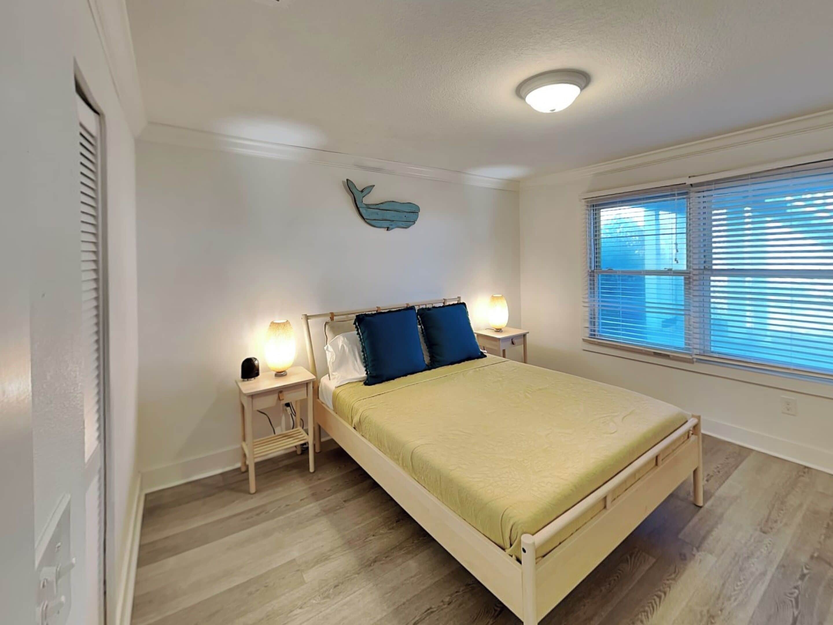 Ein Schlafzimmer mit einem Bett, zwei Nachttischlampen, einer Wandleuchte, einem Fenster mit Jalousien und einem Wandbild.